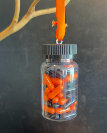 A clear plastic prescription bottle Halloween ornament with a black cap, filled with faux orange and black capsules. Pills are empty. For Decorative Use Only.