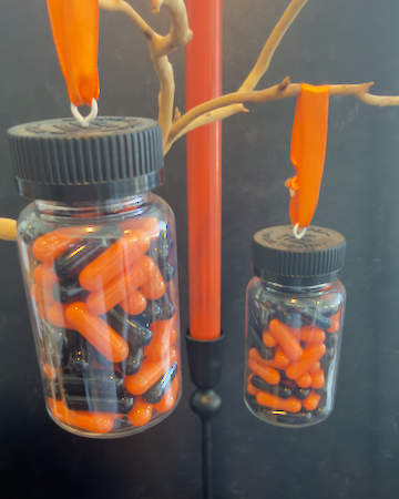 A clear plastic prescription bottle Halloween ornament with a black cap, filled with faux orange and black capsules. Pills are empty. For Decorative Use Only.