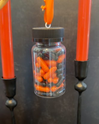 A clear plastic prescription bottle Halloween ornament with a black cap, filled with faux orange and black capsules. Pills are empty. For Decorative Use Only.