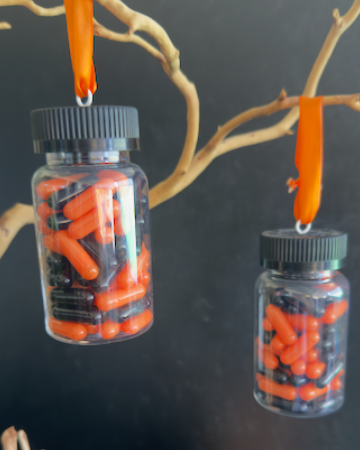 A clear plastic prescription bottle Halloween ornament with a black cap, filled with faux orange and black capsules. Pills are empty. For Decorative Use Only.