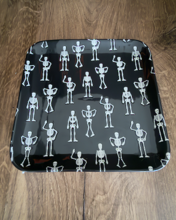 A square ceramic black appetizer tray with rounded corners, featuring a repeating pattern of small white stick-figure skeletons. Approx. 8.25" square.