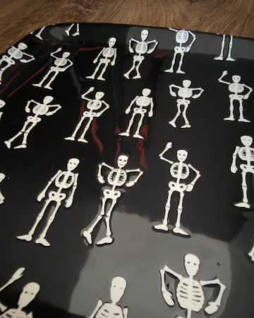 A square ceramic black appetizer tray with rounded corners, featuring a repeating pattern of small white stick-figure skeletons. Approx. 8.25" square.