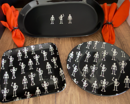 A Halloween-themed black dinnerware set on a wooden table, featuring a long oval platter, a square tray, and a round salad plate, all printed with a white skeleton figure pattern.