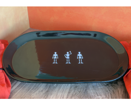 A large ceramic glossy black oval serving platter with three small silver skeleton figurines arranged in a row at the center. approx. 20" x 10"