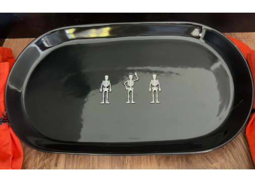 A large ceramic glossy black oval serving platter with three small silver skeleton figurines arranged in a row at the center. approx. 20" x 10"