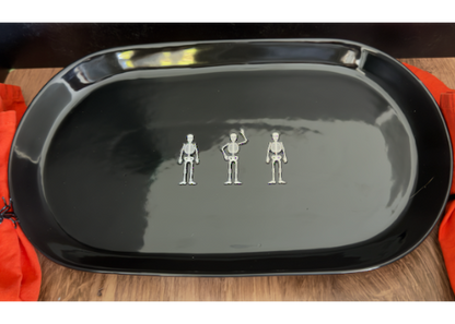 A large ceramic glossy black oval serving platter with three small silver skeleton figurines arranged in a row at the center. approx. 20" x 10"